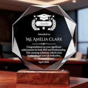 May include: A crystal award with a black face and a wooden base. The award is engraved with the name "Amelia", a graduation cap, laurel wreath, and text that reads "Awarded to MS. AMELIA CLARK" and "April 2026".