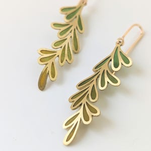 May include: A pair of gold-toned metal drop earrings shaped like fern leaves. Each earring features a delicate, open-work branch design with internal enamel inlays that transition in color from deep forest green to a lighter, golden yellow hue.