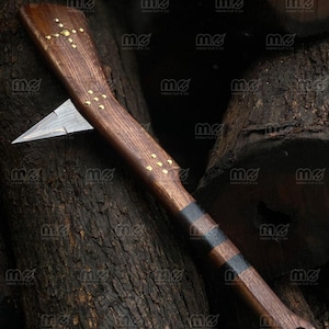 Op de afbeelding: Een handbijl met een scherp, puntig metalen blad bevestigd aan een lange, donkerbruine houten steel. De steel is versierd met ingelegde koperen pinnen in geometrische patronen en heeft afwisselende donkere houten banden bij de basis.