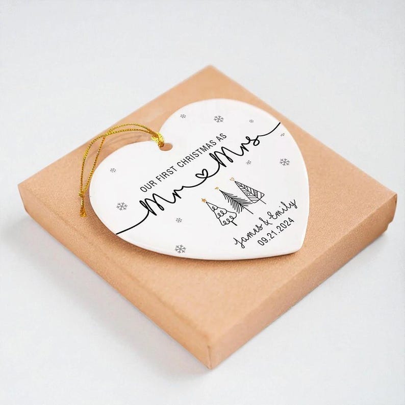 May include: A heart-shaped white ceramic Christmas ornament with gold string, resting on a brown box. The ornament features the text "Our First Christmas as Mr & Mrs", snowflakes, and two Christmas trees. The names "James & Emily" and the date "09.21.2024" are also included.