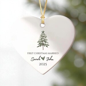 May include: A white heart-shaped Christmas ornament with a gold string. The ornament features a green watercolor pine tree and the text "FIRST CHRISTMAS MARRIED" with the names "Sarah" and "John" and the year "2025".