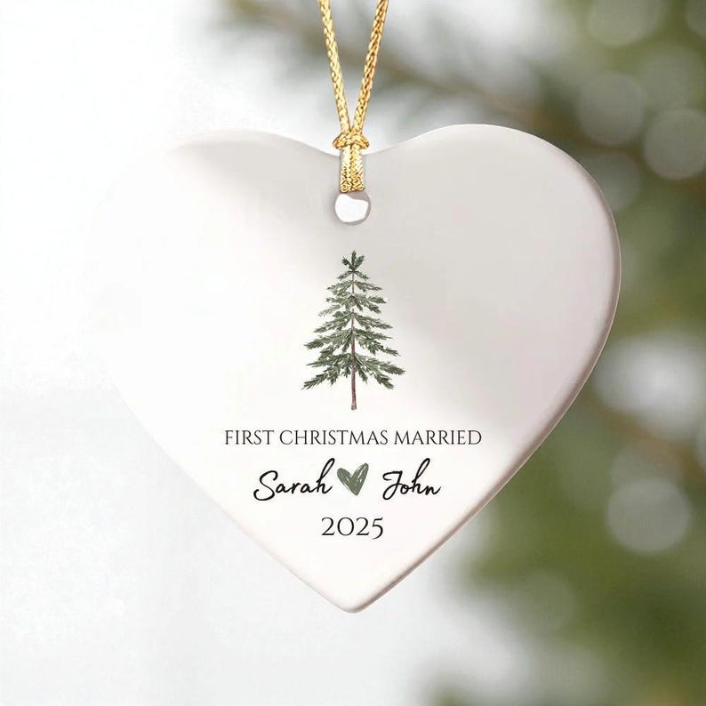 May include: A white heart-shaped Christmas ornament with a gold string. The ornament features a green watercolor pine tree and the text "FIRST CHRISTMAS MARRIED" with the names "Sarah" and "John" and the year "2025".