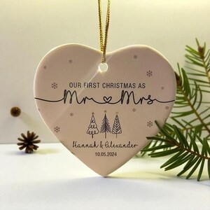 May include: A heart-shaped Christmas ornament with a gold string for hanging. The ornament is light pink and features the text "OUR FIRST CHRISTMAS AS Mr & Mrs" with a line and three Christmas trees. The names "Hannah & Alexander" and the date "10.05.2024" are also printed.