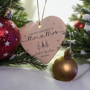 May include: A heart-shaped Christmas ornament with the text "Our First Christmas as Mr & Mrs" and the names Axel & Yarlin, with the date 08.24.2024. The ornament is light pink and decorated with small snowflakes and two Christmas trees. A red and white Christmas ball is visible in the background.
