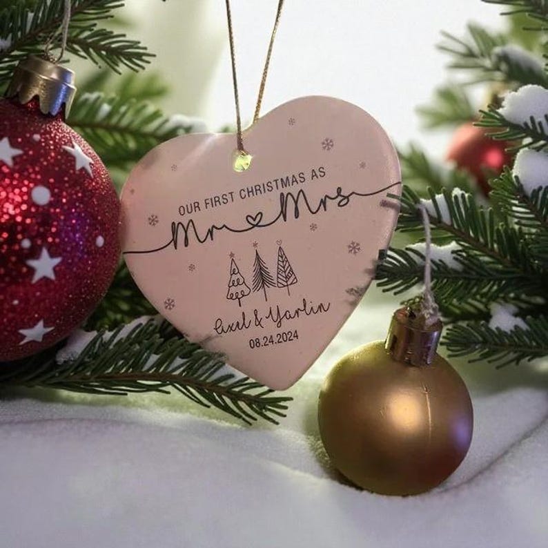 May include: A heart-shaped Christmas ornament with the text "Our First Christmas as Mr & Mrs" and the names Axel & Yarlin, with the date 08.24.2024. The ornament is light pink and decorated with small snowflakes and two Christmas trees. A red and white Christmas ball is visible in the background.