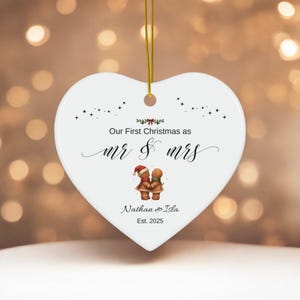 May include: Heart-shaped Christmas ornament with gold string. The white ceramic ornament features the text "Our First Christmas as mr & mrs" and the names "Nathan & Isla Est. 2025". Two gingerbread people are also depicted.