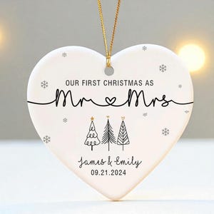 May include: A white heart-shaped Christmas ornament with gold string. The ornament reads "OUR FIRST CHRISTMAS AS Mr & Mrs" with a heart symbol. Below are three Christmas trees and the names "James & Emily" with the date "09.21.2024".