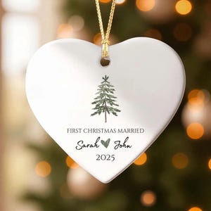 May include: White heart-shaped Christmas ornament with a gold string. The ornament features a green Christmas tree design and the text "FIRST CHRISTMAS MARRIED" with the names "Sarah &hearts; John" and the year "2025".