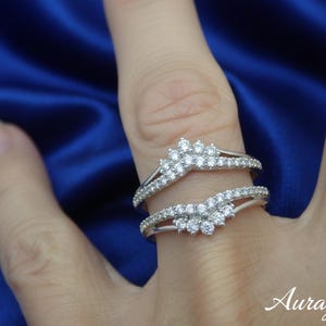 May include: Two silver rings with small, clear gemstones. The rings are arranged to appear as a set, with a curved design. The background is a deep blue fabric. The word "Auragrace" is in the bottom right corner.