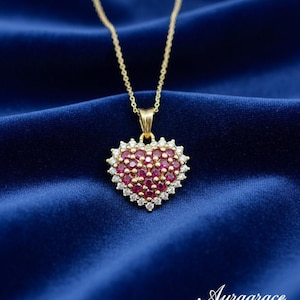 May include: A gold necklace featuring a heart-shaped pendant. The pendant is set with small, round, red gemstones at the centre, encircled by a border of clear, sparkling stones. The necklace is displayed against a deep blue fabric.