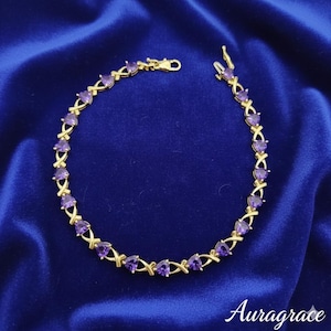 May include: Gold-toned bracelet featuring purple heart-shaped gemstones. The bracelet has an X-shaped design between each gemstone, and a clasp closure. The bracelet is displayed on a blue satin surface. The word "Auragrace" is written in cursive at the bottom.