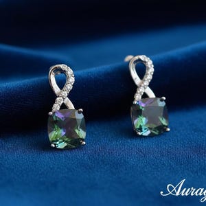 May include: A pair of silver-toned earrings with a square, multi-colored gemstone. The top of each earring has a loop design encrusted with small, clear stones. The background is a deep blue fabric. The word "Auragrace" is in the bottom right corner.