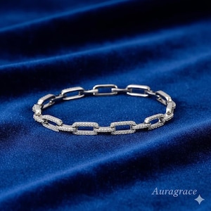 May include: A silver link bracelet with rectangular links, each set with small, sparkling stones. The bracelet is shown on a deep blue velvet surface, which contrasts with the jewellery's design. The brand name "Auragrace" is visible.