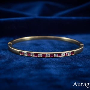 May include: A gold-toned bangle bracelet with a row of alternating red and clear square-cut gemstones. The bracelet is set against a deep blue velvet background. The word "Auragrace" is in the bottom right corner.