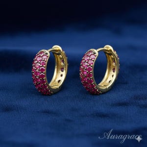 Puede incluir: Un par de pendientes de aro dorados, incrustados con pequeñas gemas redondas de color rojo rubí. Los pendientes se presentan sobre un fondo azul oscuro, con el nombre de la marca "Auragrace" en la esquina inferior derecha.