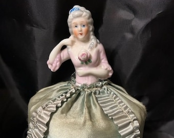 May include: A vintage porcelain doll pin cushion. The doll has a pink bodice and a blue bow in her hair. The pin cushion is a light green color with a ruffled edge and decorative trim.
