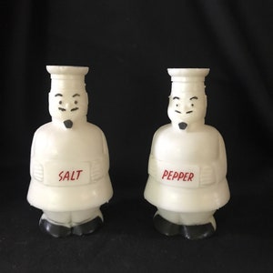 May include: White ceramic salt and pepper shakers shaped like chefs with black eyes and eyebrows. The shakers have red text that says "SALT" and "PEPPER".