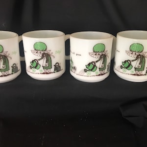 May include: Set of four white ceramic mugs with a green and brown illustration of a girl watering flowers. The mugs have the text "Let's all grow" on the front.