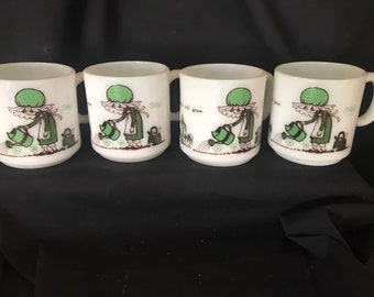 May include: Set of four white ceramic mugs with a green and brown illustration of a girl watering flowers. The mugs have the text "Let's all grow" on the front.