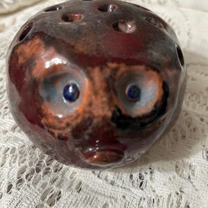 May include: A ceramic, spherical object with a face-like design. The object is primarily brown with reddish-brown and orange accents. It has two blue eyes and several holes on top, possibly for holding flowers. The object is on a white lace surface.