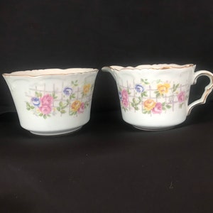 Puede incluir: Un juego de jarra de crema y azucarero de porcelana blanca con un patrón floral. El patrón presenta rosas rosas, amarillas y azules con hojas verdes sobre un fondo blanco. La jarra tiene un asa y el azucarero es redondo.