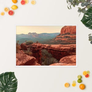 May include: Framed landscape photograph of a natural red rock arch in a desert setting. The image shows the arch's distinctive shape, with distant mountains and a clear sky in the background. The photograph is presented in a white frame.