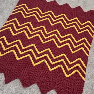 May include: A handmade crochet blanket featuring a chevron pattern. The blanket has a deep burgundy base color with bright yellow zig-zag stripes. The edges of the blanket are scalloped.