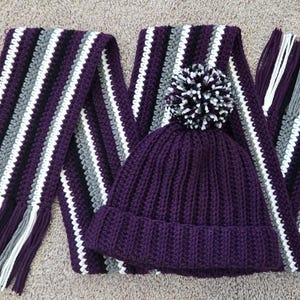 May include: A knitted scarf and hat set in shades of purple, white, gray, and black. The scarf has vertical stripes and fringe. The hat is solid purple with a white and black pom-pom. This cozy set is perfect for cold weather.
