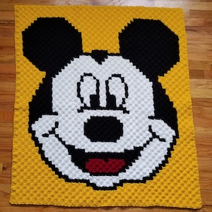 Può includere: Una coperta all'uncinetto con uno sfondo giallo e un viso di Topolino bianco e nero al centro. Topolino sorride con le sue iconiche orecchie nere.