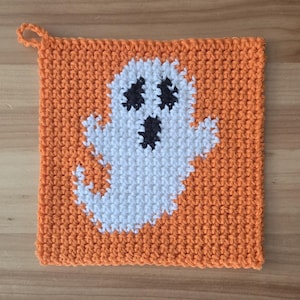 May include: A square crochet potholder in bright orange yarn featuring a centred white ghost motif with black yarn details for the eyes and mouth. The potholder has a small loop at the top left corner for hanging and a textured stitch pattern.