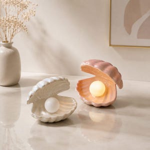 May include: Two ceramic clam shell lamps with glossy finishes. One shell is white with a white sphere light, and the other is a soft pink with a warm glowing sphere light. Both shells are open, displaying the spherical light bulbs inside.