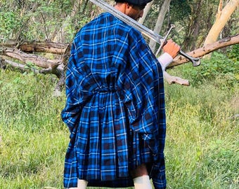 May include: A person wearing a blue and black plaid kilt and cape, black beret, white socks, and black boots, holding a long silver sword. The outfit is set against a backdrop of green grass and trees.