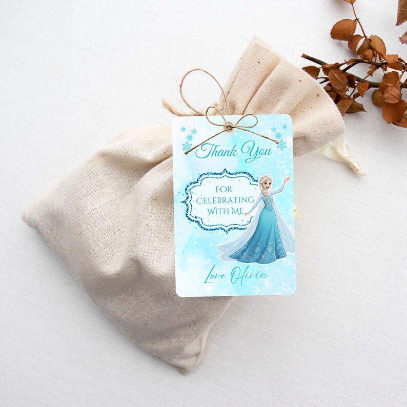 Puede incluir: Una bolsa de regalo de tela beige claro con una etiqueta atada con hilo. La etiqueta presenta una imagen de dibujos animados de una princesa con un vestido azul, con las palabras "Thank You" y "For Celebrating With Me". La etiqueta est&aacute; personalizada con el nombre "Olivia".