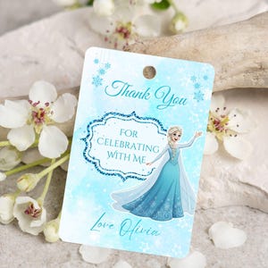 Puede incluir: Una etiqueta de regalo azul claro con el texto "Thank You For Celebrating With Me Love Olivia". La etiqueta presenta una ilustraci&oacute;n de dibujos animados de una princesa con un vestido azul, con un dise&ntilde;o de copo de nieve.