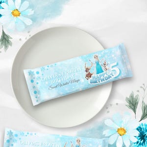 Puede incluir: Un envoltorio de barra de caramelo azul claro con el texto "Olivia's Frozen Birthday Sweet Winter Magic" y un gr&aacute;fico de Elsa, Olaf y Sven. El envoltorio tambi&eacute;n tiene el n&uacute;mero 5 y las palabras "Happy Birthday". La barra de caramelo est&aacute; en un plato blanco.