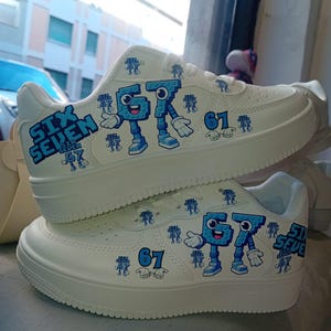 May include: White sneakers with blue cartoon graphics. The sneakers feature the text "SIX SEVEN" and the number "61" in blue. The cartoon graphics are of a robot-like character. The shoes are stacked on top of each other.