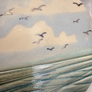 May include: A semi-circular ceramic wall plaque featuring a textured, crackled glaze in shades of light blue and cream. The surface depicts a coastal scene with stylised dark blue birds flying across a cloudy sky above rippled ocean waves.