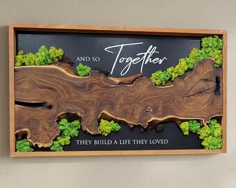 May include: A framed wooden wall art piece with a dark background. The art features a natural wood grain design, green moss accents, and the words "And So Together They Build a Life They Loved" in white script.