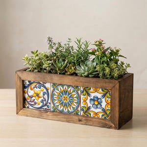 May include: A rectangular wooden planter box filled with various green succulents and flowering plants. The front of the box features three decorative tiles with colorful floral patterns in yellow, blue, and white.