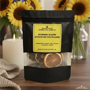 May include: A black pouch of "Summer Blend Stovetop Potpourri" with a yellow label. The label lists ingredients: lemon, lime, orange, lavender, and ginger. The pouch contains dried citrus slices and is displayed with sunflowers and a lit candle.