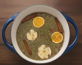 May include: A blue and white enamel pot filled with rosemary, orange slices, apple slices, and cinnamon sticks. The pot sits on a wooden surface, creating a warm and inviting aesthetic. The ingredients suggest a seasonal or aromatic preparation.