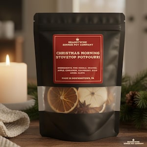 May include: A black pouch of Christmas Morning stovetop potpourri with a clear window showing dried orange slices, star anise, and apple slices. The label reads "Christmas Morning Stovetop Potpourri" and lists ingredients. A lit candle and greenery are in the background.