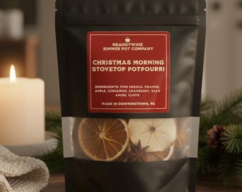 May include: A black pouch of Christmas Morning stovetop potpourri with a clear window showing dried orange slices, star anise, and apple slices. The label reads "Christmas Morning Stovetop Potpourri" and lists ingredients. A lit candle and greenery are in the background.
