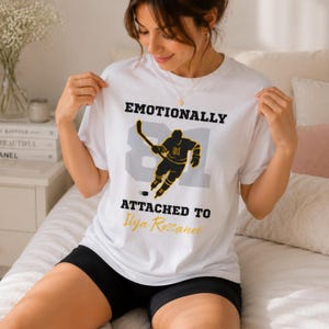 May include: White t-shirt with the text "Emotionally Attached To Ilya Rozanov". The shirt features a hockey player graphic in black and gold with the number 81. The shirt is worn by a person.