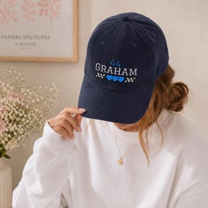 May include: Navy blue corduroy baseball cap with embroidered text "44 GRAHAM" in white and blue. The design includes blue hearts and checkered flag accents. A stylish accessory for casual wear.