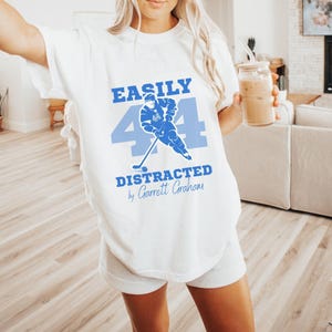 White tee blue Easily Distracted Garrett Graham 44 hockey print, blonde model holding iced latte living room