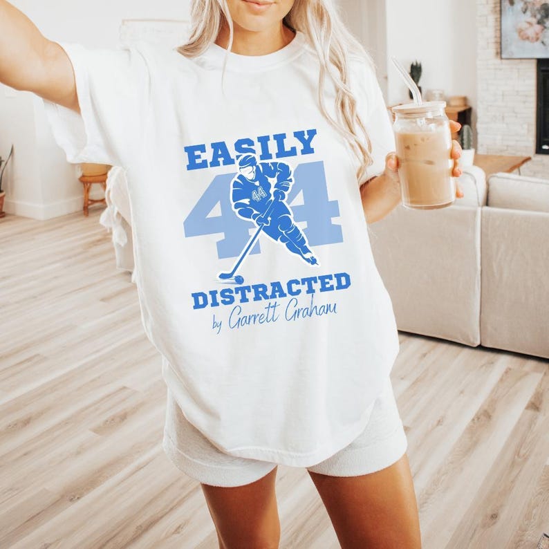 White tee blue Easily Distracted Garrett Graham 44 hockey print, blonde model holding iced latte living room