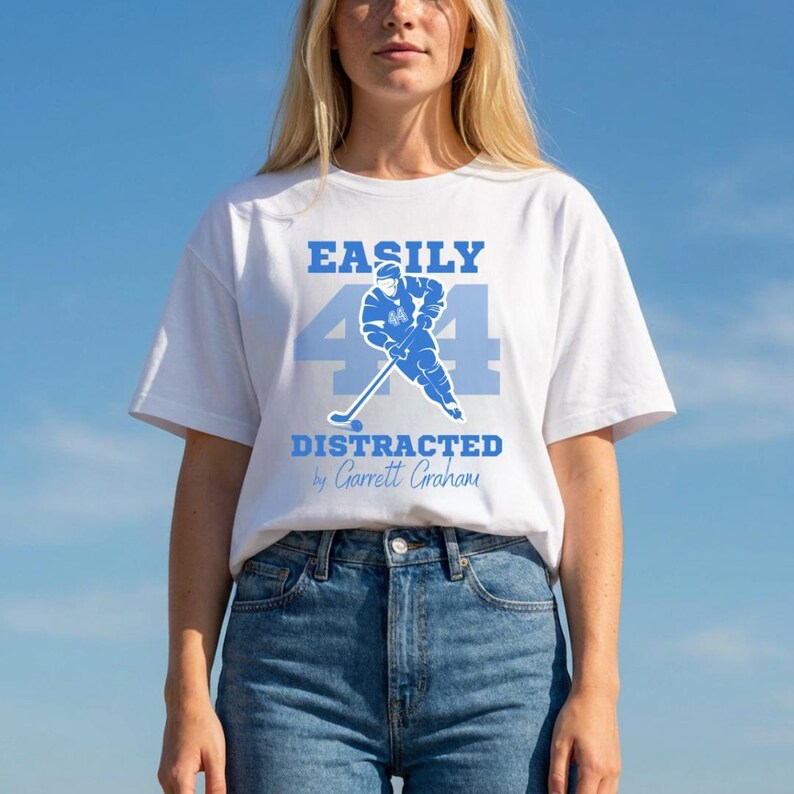 White tee blue Easily Distracted by Garrett Graham 44 hockey player graphic, blonde standing model blue sky