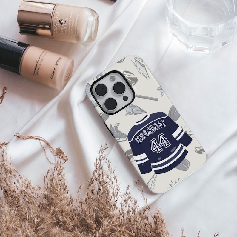 Cream iPhone Pro Graham 44 navy hockey jersey case flat lay on white silk with makeup and dried grasses