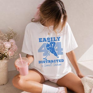 Op de afbeelding: Wit T-shirt met een blauwe print met de tekst "EASILY 44 DISTRACTED by Garrett Graham". De print toont een hockeyspeler en het nummer 44. Het shirt heeft een casual, oversized pasvorm.